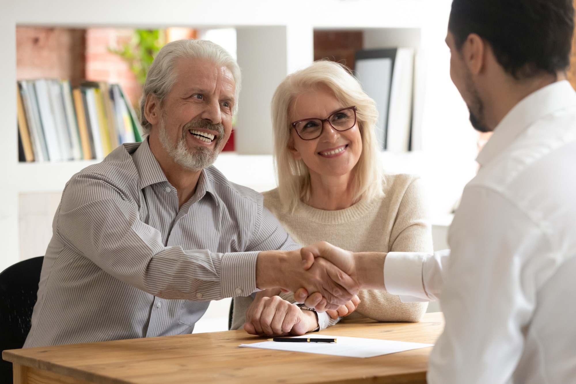 An older couple smiles and shakes hands with a professional man at a table, suggesting a successful agreement or partnership. Papers and a pen are visible.