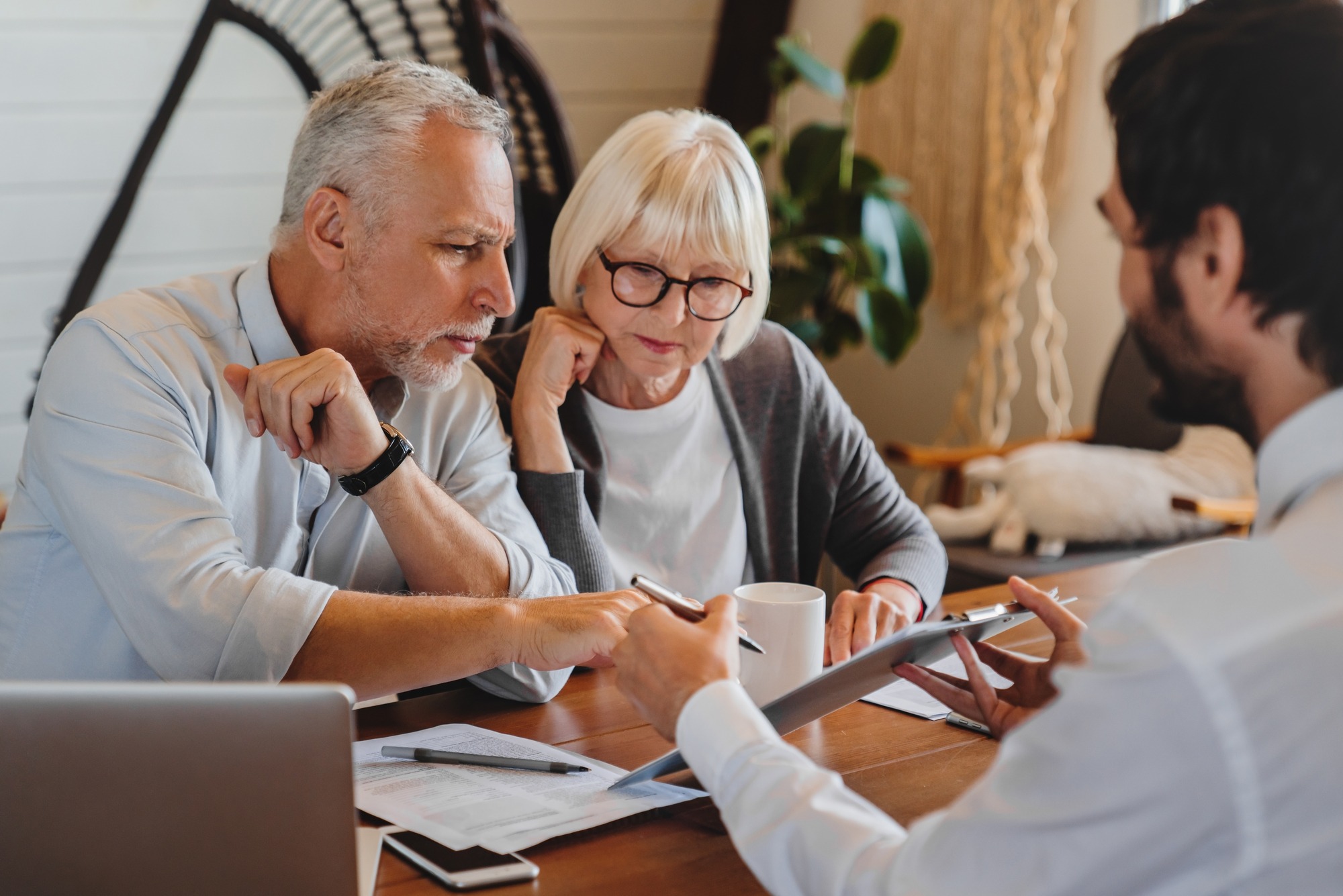 A senior couple engages in a discussion with a consultant over a tablet, examining documents on a table in a cozy, modern office setting.